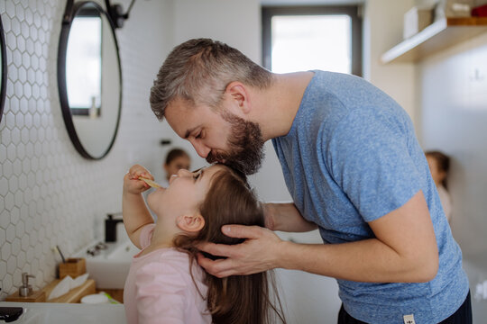 Father kissing his daughter while she is brushing her teeth in bathroom, morning routine concept.