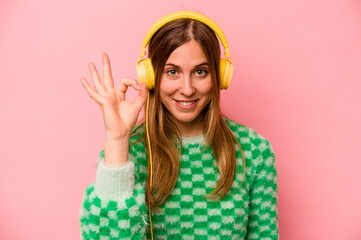 Young caucasian woman listening to music isolated on pink background cheerful and confident showing ok gesture.