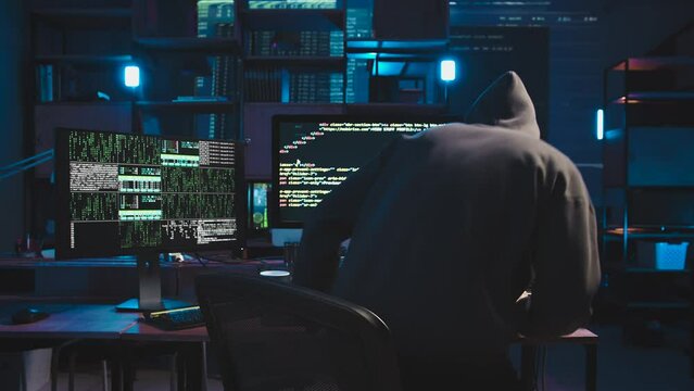 A gloomy and secretive programmer comes and sits down at the computer desk. In front of him are two monitors and a laptop. He writes complex multi-level code for the program. Room at dusk
