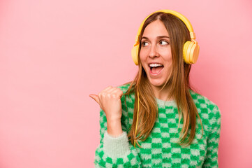 Young caucasian woman listening to music isolated on pink background points with thumb finger away, laughing and carefree.