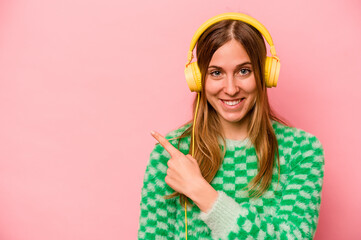 Young caucasian woman listening to music isolated on pink background smiling and pointing aside, showing something at blank space.