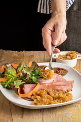 View of woman's hand putting caramelized onions in a plate of pastrami with salad, on wooden table, black background, vertical