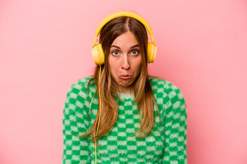 Young caucasian woman listening to music isolated on pink background shrugs shoulders and open eyes confused.