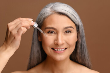 Beautiful middle aged Asian woman applies regenerating serum on the face. Charming grey-haired 50s...