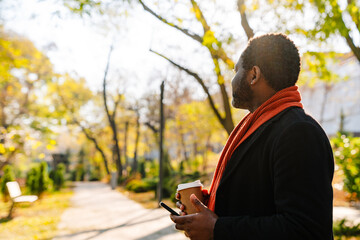 Black man using cellphone and drinking coffee while strolling in park