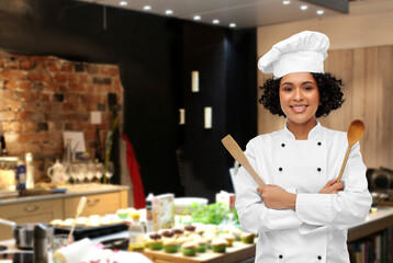 cooking, culinary and people concept - happy smiling female chef in toque with wooden spoon and spatula over restaurant kitchen background