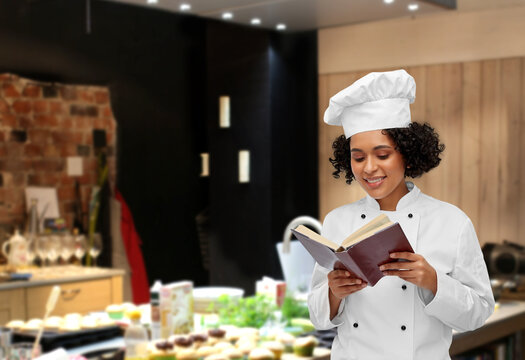 cooking, culinary and people concept - happy smiling female chef in white toque and jacket reading cook book over restaurant kitchen background