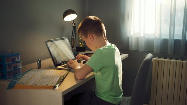 Boy sitting at the desk doing homework at home using laptop.