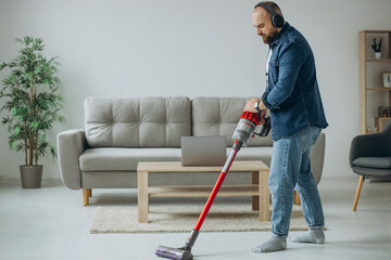 Man doing house work with accumulator vacuum cleaner and listening to music on headphone