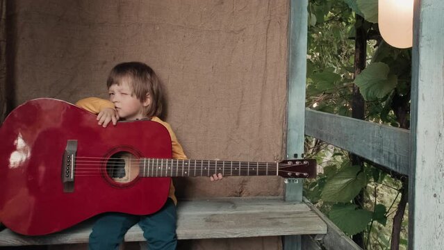 Small, funny child with a large acoustic guitar is sitting on the wooden porch of a country house. The concept of humor and laughter