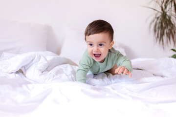 cheerful baby in a green cotton bodysuit is lying on his stomach on a white bed