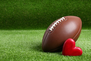 American football ball and heart on green grass. Space for text