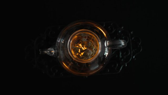 Still life. Tea ceremony. Brewing herbal tea in a beautiful glass teapot. Pour boiling water in a thin stream. A small candle warms up the kettle. Pleasant lighting and reflections in the dishes.