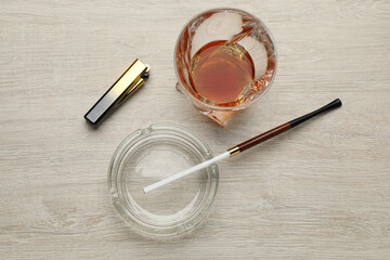 Glass ashtray with long cigarettes holder, lighter and alcohol drink on wooden table, flat lay