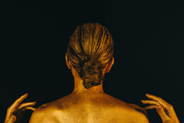 A young woman covered in gold paint stands with her back against a black background. Topics of aesthetics, fashion, design, creativity, beauty, style, hair, makeup, body painting