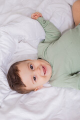 cute smiling baby in a green cotton bodysuit is lying on a white bed on his back. top view
