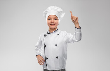cooking, culinary and profession concept - happy smiling little girl in chef's toque and jacket pointing finger up over grey background