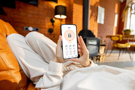Woman using phone with running smart home security application, while lying on a couch at home. Image focused on device screen