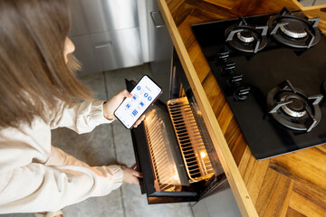 Woman holding phone with running smart home application, controlling kitchen devices remotely. Smart kitchen appliances concept