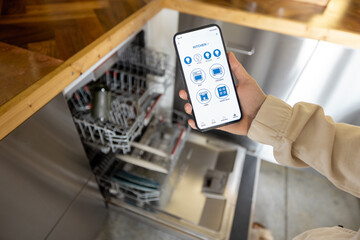 Woman holding phone with running smart home application, controlling dishwasher remotely. Smart...