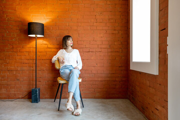 Full length portrait of a young woman sits on chair near floor lamp on brick wall background at home - obrazy, fototapety, plakaty