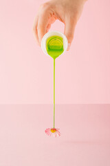 A creative spring and flower arrangement made by a hand that pours green color and blends with the stem of a daisy. Minimal collage concept on pink background. Plastic and nature.
