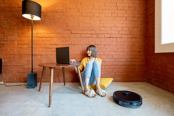 Young woman work on laptop computer, sitting on floor while robotic vacuum cleaner cleaning floor...