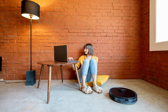 Young woman work on laptop computer, sitting on floor while robotic vacuum cleaner cleaning floor at home. Concept of smart home gadgets and work from home