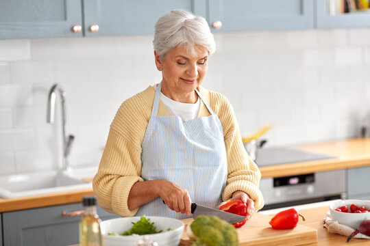 healthy eating, food cooking and culinary concept - happy smiling senior woman with knife chopping red pepper on kitchen at home