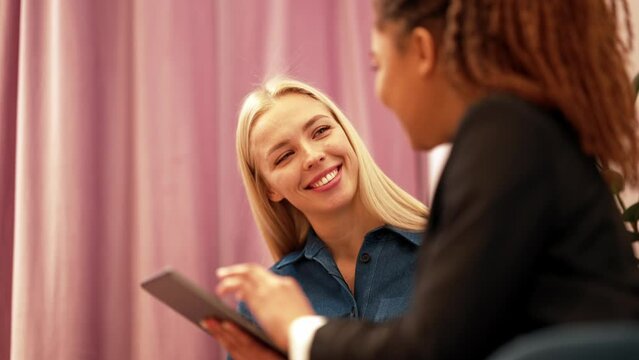 Smiling African saleswoman looking at the tablet with blonde client in the fitting room in a clothing store