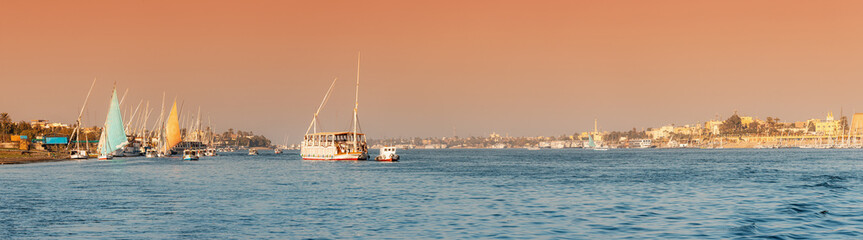 Fototapeta premium Panoramic view of traditional Egyptian ferry boats and cruise ships down the Nile with ancient temples of Luxor in the background