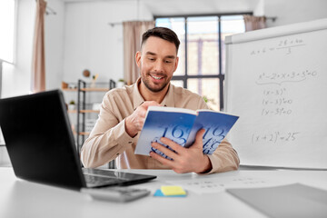 distant education, school and remote job concept - happy smiling male math teacher with laptop computer, book and flip chart having online class or video call at home office