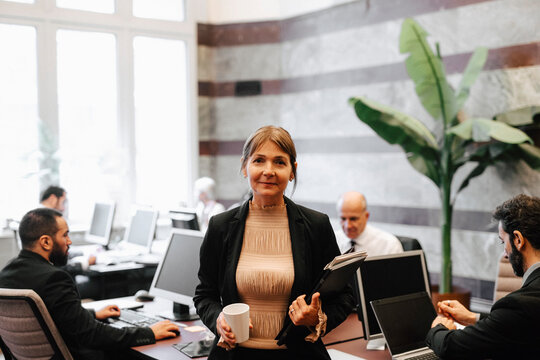 Portrait of smiling mature female leader holding coffee cup and documents at law office