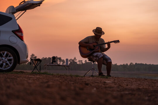 An Asian man sits in the back of his car while playing a guitar in a small campsite at sunset.