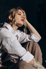 Portrait of a fashionable retro girl in a white shirt, suspenders and brown wide leg pants, sitting on the floor in the sun in the room
