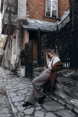 Fotobehang A posing beautiful girl with blond hair stands in the old town near a beautiful door on the street in brown trousers, a brown coat, a white blouse, black shorts and a hat © Илля Червонящий