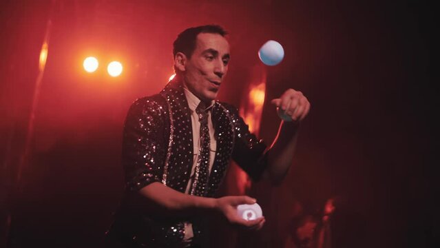 A jolly man in a circus professionally juggles three luminous balls in beautiful lighting, slow motion
