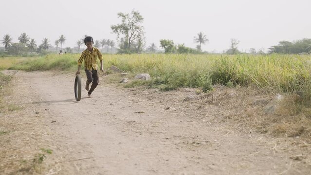 Village boy kid playing with tyre wheel rolling at at near paddy field- concept of holidays, freedom, poverty and childhood lifestyle