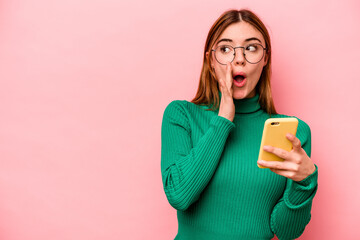 Young caucasian woman holding mobile phone isolated on pink background is saying a secret hot braking news and looking aside - obrazy, fototapety, plakaty