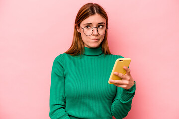 Young caucasian woman holding mobile phone isolated on pink background confused, feels doubtful and...