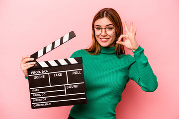 Young caucasian woman holding a clapperboard isolated on pink background cheerful and confident...