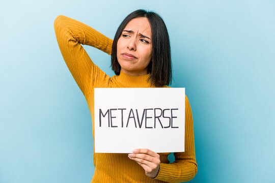 Young hispanic woman holding metaverse placard isolated on blue background touching back of head, thinking and making a choice.