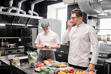 Young chef in uniform checking freshness of vegetables standing near kitchen table with his assistant making notes behind him