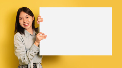 Charming young Asian woman holding blank paper poster with mockup, smiling at camera on yellow...