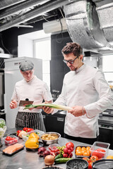 Young chef in uniform standing in kitchen and checking quality of fresh vegetables on table before cooking