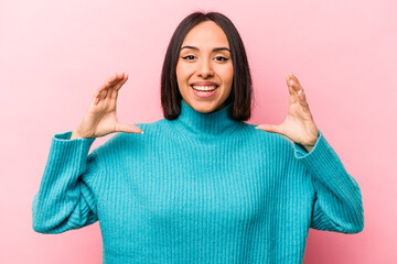 Young hispanic woman isolated on pink background holding something with palms, offering to camera.