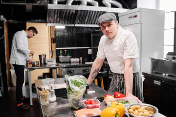 Portrait of young cook in uniform looking at camera while standing at table with fresh vegetables and cooking in kitchen