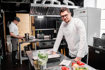 Portrait of young chef in uniform looking at camera while standing at his workplace with food ingredients during work in kitchen