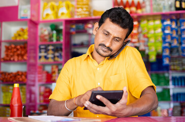 Kirana or grocery marchant calculating order price while taking on mobile phone from customer -...