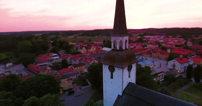 Aerial Panning Shot Of Clock On Tall Church Amidst Houses In Town By Sea At Sunset - Mariefred, Sweden